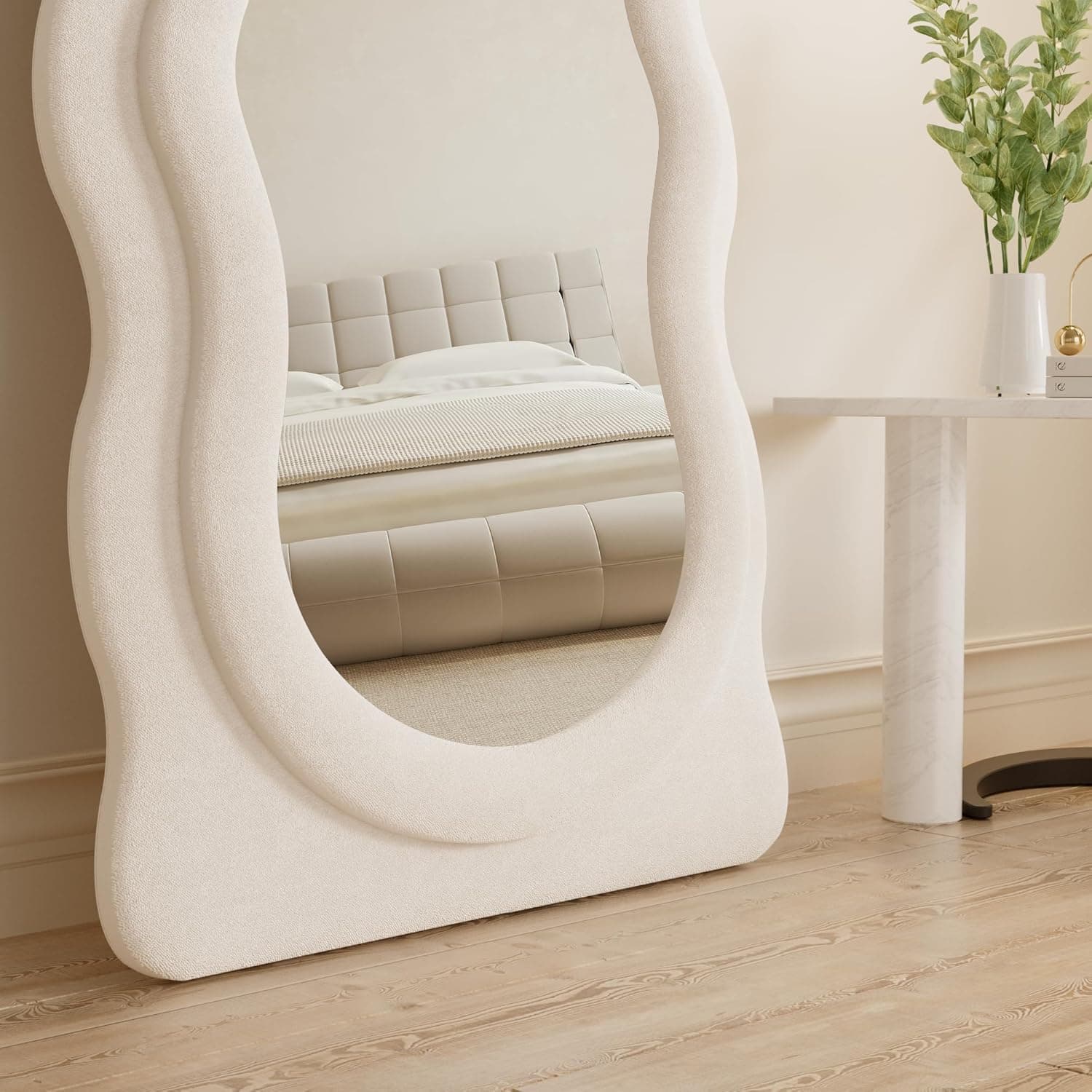 Elegant beige upholstered bed with a tufted headboard, reflected in a stylish, sculptural full-length mirror in a modern, minimalist bedroom setting.