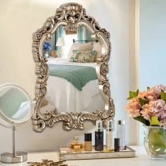 Elegant ornate silver mirror reflecting a cozy bedroom with plush pillows and a teal blanket. Perfect for upscale home décor and bedroom styling.