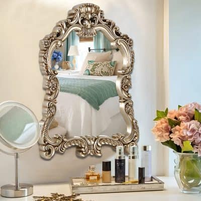 Elegant ornate silver mirror reflecting a cozy bedroom with plush pillows and a teal blanket. Perfect for upscale home décor and bedroom styling.