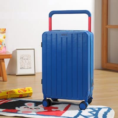 Blue hard shell travel suitcase with red handle and four spinner wheels for easy mobility on wooden floor with colorful rug and decorative framed artwork in background.