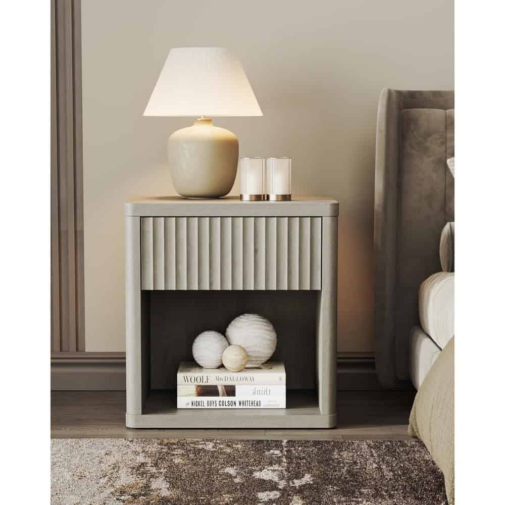 Modern beige bedside table with ribbed drawer front and lower shelf holding decorative books and spheres, placed beside a plush bed with taupe upholstery and a soft beige bedding, illuminated by a stylish table lamp.