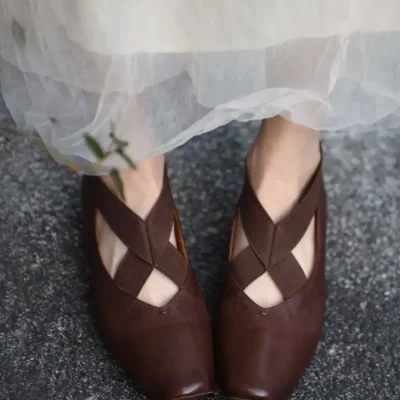 Brown vintage-style leather ballet flats with crisscross straps on the toes, worn with a light, sheer skirt; stylish footwear for everyday and special occasions.