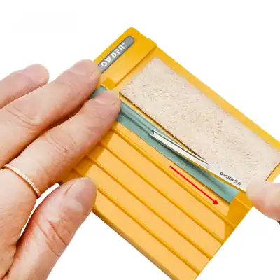 Abrasive testing block for dental and optical lens polishing or blade edge testing, bright yellow plastic with embedded abrasive surface and metal tool inside for precise sharpening or grinding use.