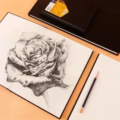 A detailed pencil sketch of a blooming rose on white paper, showcasing realistic shading and intricate petal details, laid out on a desk with a black notebook and a pencil.