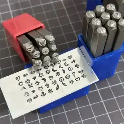 Stamped metal alphabet and symbol stamps for jewelry and crafts, displayed in red and blue plastic cases, used for creating custom designs on metal surfaces.