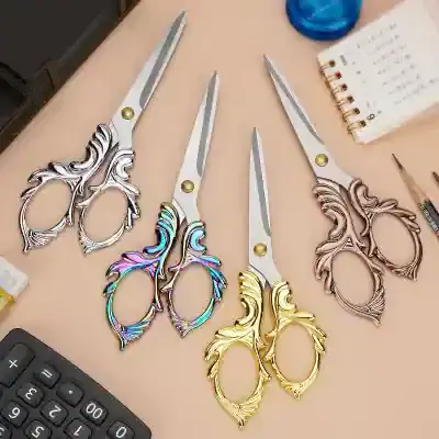Scissor manicure tools with ornate handles in silver, rainbow, gold, and rose gold finishes for beauty and grooming, displayed on a beige table with notebook and calculator in the background.