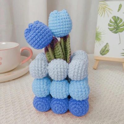 Colorful crocheted stuffed flowers in shades of blue, purple, and gray, perfect for home decor or gifts, displayed on a table with a tea cup and botanical artwork in the background.