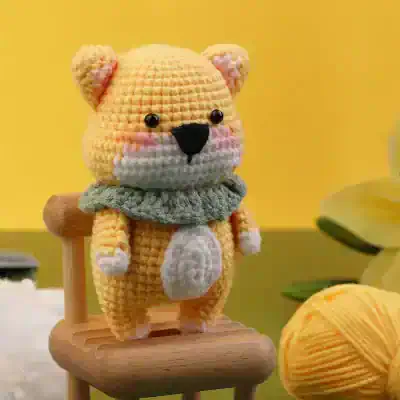 Soft yellow crochet teddy bear with black eyes and nose, wearing a gray ruffled collar, sitting on a small wooden chair with a yellow background.