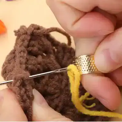 Knitting close-up, hand stitching brown fuzzy yarn with yellow thread, gold textured ring on finger, craft sewing project, homemade yarn toy, detailed view of knitting process, creative knitting and handiwork, craft hobby and textile art, DIY knitting, fiber arts and needlework.