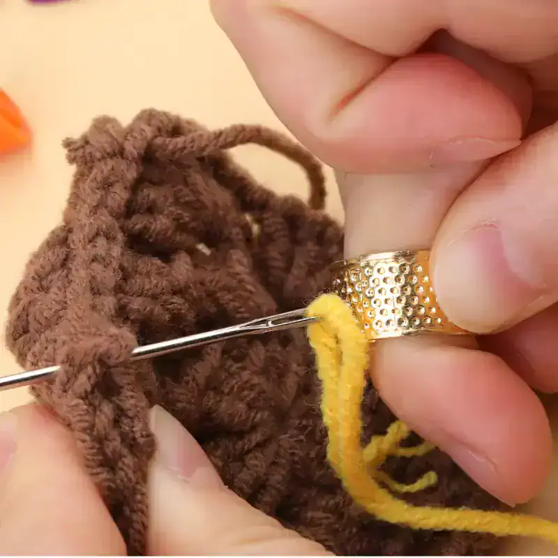 Knitting close-up, hand stitching brown fuzzy yarn with yellow thread, gold textured ring on finger, craft sewing project, homemade yarn toy, detailed view of knitting process, creative knitting and handiwork, craft hobby and textile art, DIY knitting, fiber arts and needlework.