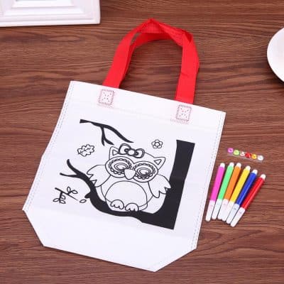 Black and white kids' coloring tote bag featuring a cartoon owl design, accompanied by colorful markers, on a wooden surface. Perfect for children's art activities, school projects, or creative play.