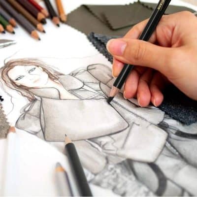 A hand sketching a fashionable woman in a stylish outfit with colored pencils and fabric swatches on a design table, illustrating fashion design and embroidery at Monalisa Store for custom clothing and tailoring services.