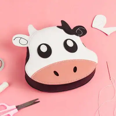 Vegan cow-shaped purse for kids on pink background with sewing tools and craft supplies, perfect for children’s accessories or playful fashion accessories.