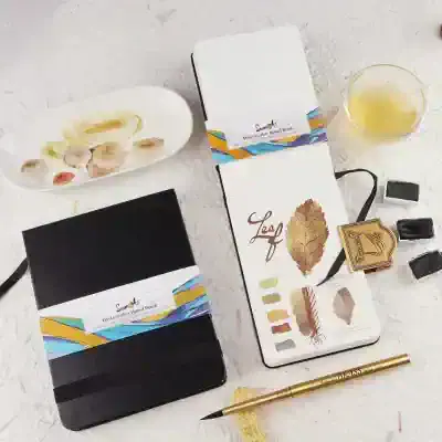 Vivid watercolor hand book, with botanical illustrations and color swatches, ideal for art and design projects, displayed alongside ink stamps, a pen, and a glass of yellow beverage.