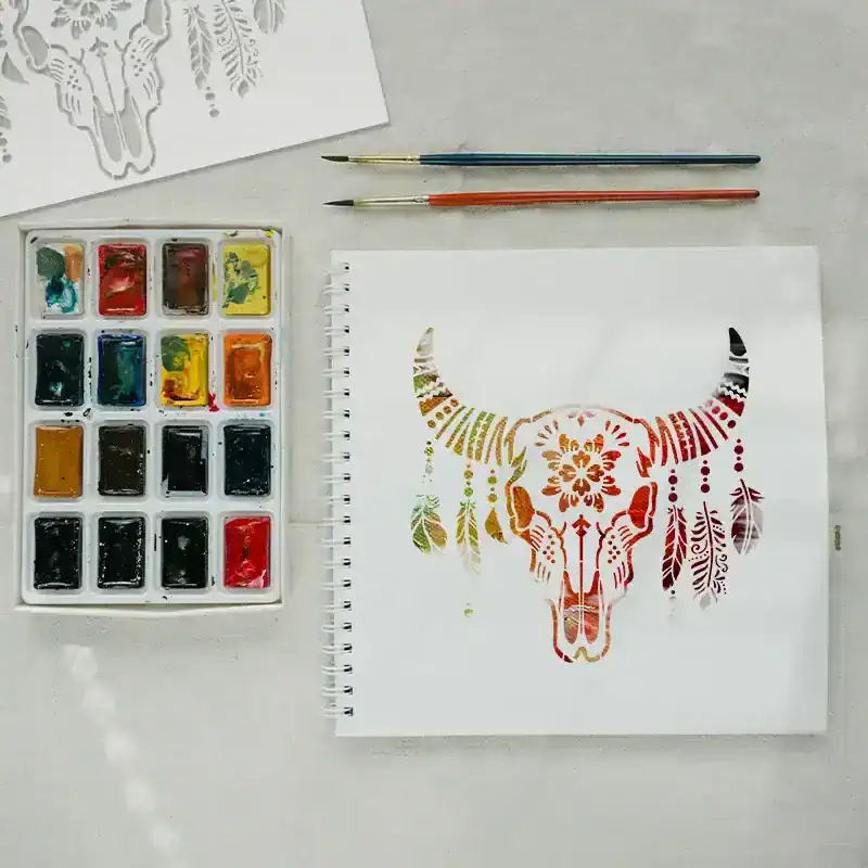Vibrant watercolor paints, a notebook with a colorful painted buffalo skull design, and fine brushes on a light gray surface, showcasing arts, crafts, and creative design inspiration.