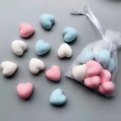 Colorful heart-shaped candies in pastel pink, white, and blue scattered on a gray surface with a transparent gift bag. Perfect for Valentine's Day, gifts, or celebrations.