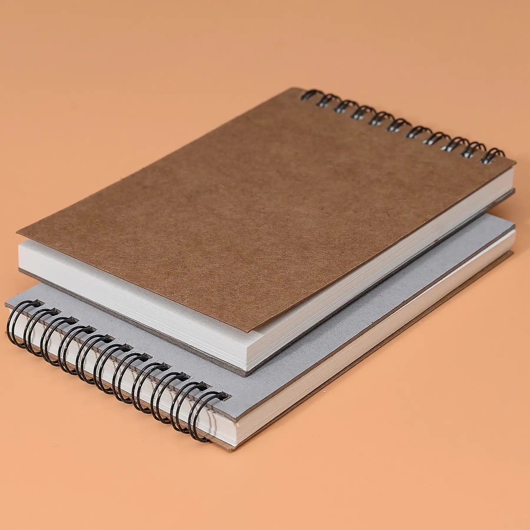 Spiral notebooks with brown, white, and gray covers on a peach background, symbolizing organization and note-taking for those interested in optical and eyewear solutions.
