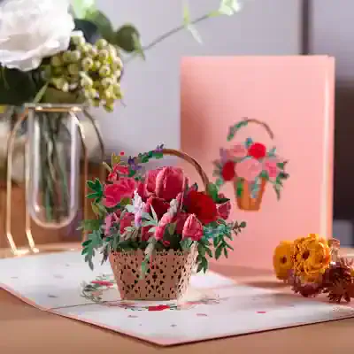 Colorful artificial flower arrangement in a decorative basket on a pink floral-themed table mat, with a flower-themed greeting card, fresh flowers in a vase, and a pink background.