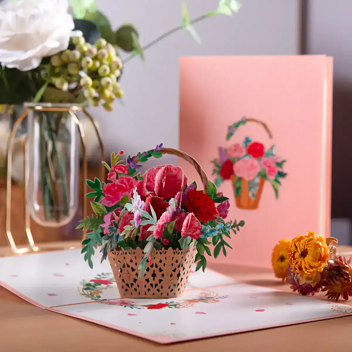Colorful artificial flower arrangement in a decorative basket on a pink floral-themed table mat, with a flower-themed greeting card, fresh flowers in a vase, and a pink background.