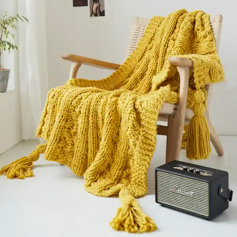 Luxurious yellow chunky knit throw blanket draped over wooden armchair in cozy home setting with vintage-inspired portable radio nearby, perfect for relaxing spaces.