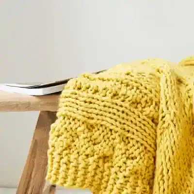Thick yellow knitted blanket draped over a wooden surface near a desk with papers, cozy home decor, soft texture, warm ambiance, perfect for bedroom or living room.
