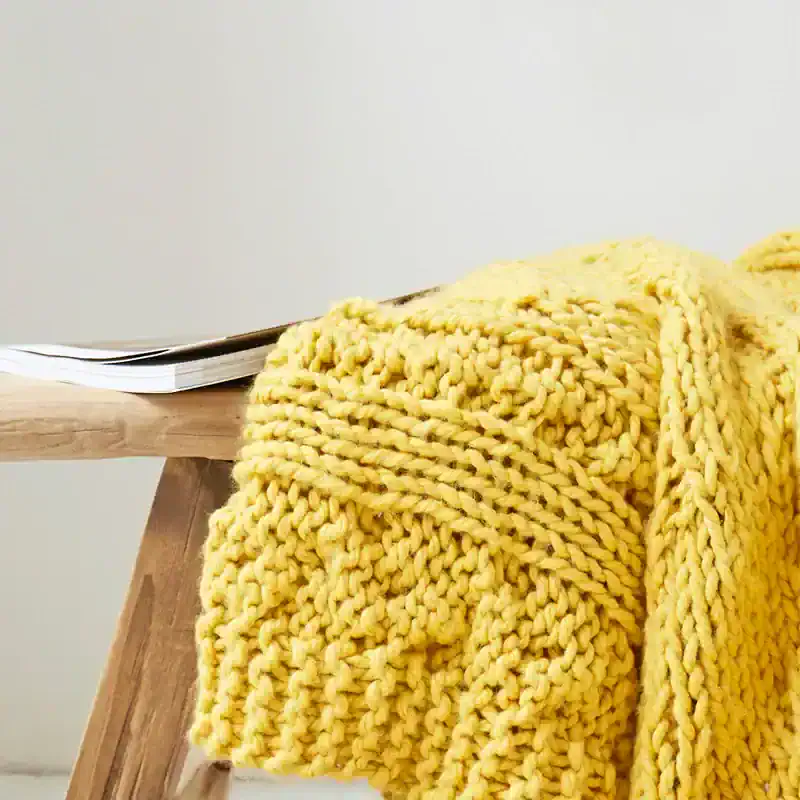 Thick yellow knitted blanket draped over a wooden surface near a desk with papers, cozy home decor, soft texture, warm ambiance, perfect for bedroom or living room.