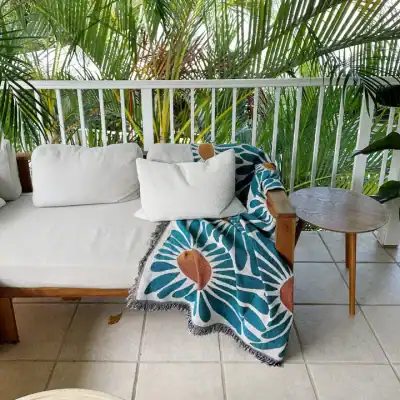 Bright outdoor balcony with tropical plants, cozy white sofa, colorful floral throw blanket, and wooden side table, perfect for relaxing and enjoying nature.