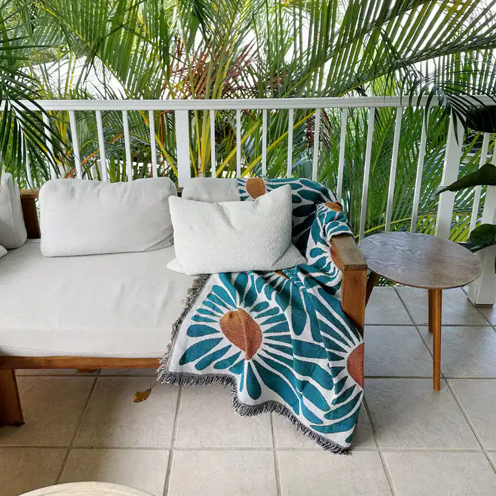 Bright outdoor balcony with tropical plants, cozy white sofa, colorful floral throw blanket, and wooden side table, perfect for relaxing and enjoying nature.