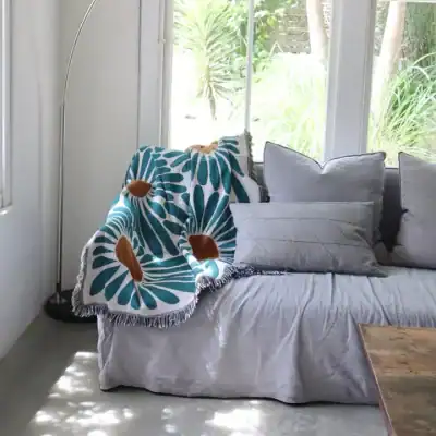 Plush sofa with decorative cushions and a colorful throw blanket in a bright, airy living room with large windows and greenery outside.