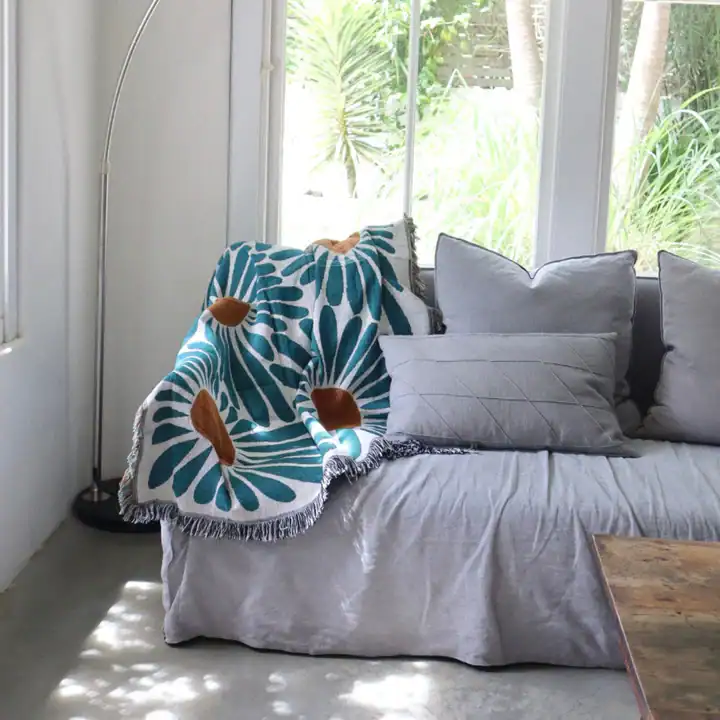 Plush sofa with decorative cushions and a colorful throw blanket in a bright, airy living room with large windows and greenery outside.