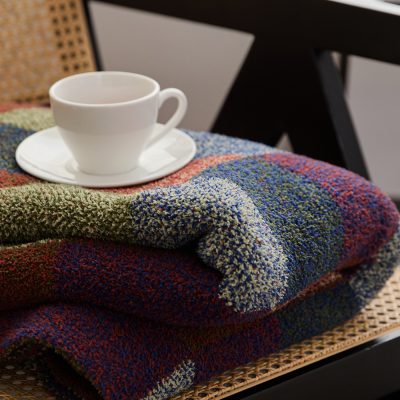 Colorful textured blanket neatly folded on a wooden chair with a white coffee cup and saucer on top for cozy, stylish home decor.