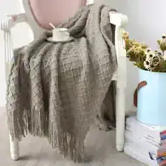 Soft beige knitted throw blanket draped over a white chair with a coffee cup, plate, and spoon on top, beside a potted daisy flower arrangement and stacked magazines, creating a cozy, stylish living space.
