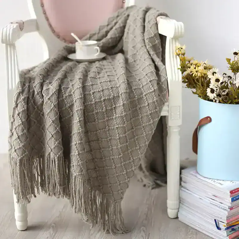 Soft beige knitted throw blanket draped over a white chair with a coffee cup, plate, and spoon on top, beside a potted daisy flower arrangement and stacked magazines, creating a cozy, stylish living space.