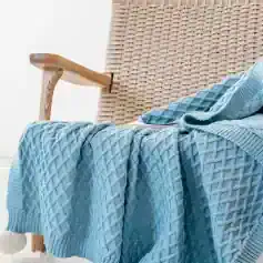Luxurious light blue knitted blanket draped over a woven chair with wooden armrest, cozy and stylish home decor for comfort and relaxation.