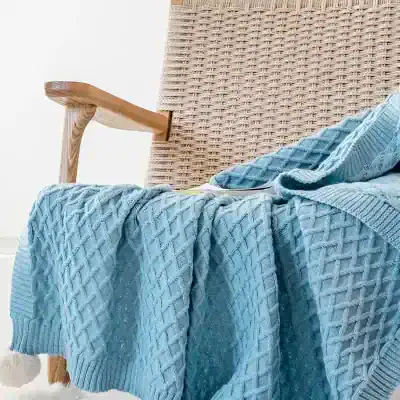 Luxurious light blue knitted blanket draped over a woven chair with wooden armrest, cozy and stylish home decor for comfort and relaxation.