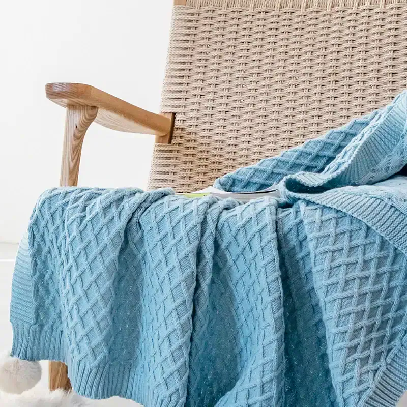 Luxurious light blue knitted blanket draped over a woven chair with wooden armrest, cozy and stylish home decor for comfort and relaxation.