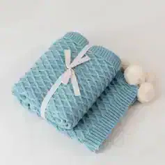 Soft light blue knitted blanket with white pom-poms, elegantly folded and tied with a satin ribbon, perfect for baby or home decor.