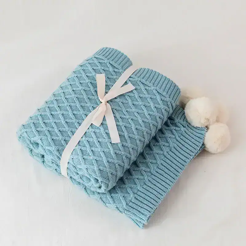 Soft light blue knitted blanket with white pom-poms, elegantly folded and tied with a satin ribbon, perfect for baby or home decor.