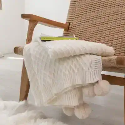 Soft cream cable-knit blanket draped over a wooden armchair in a cozy, modern space. The blanket features pom-pom tassels, perfect for adding warmth and style to your home decor.