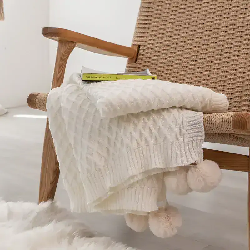 Soft cream cable-knit blanket draped over a wooden armchair in a cozy, modern space. The blanket features pom-pom tassels, perfect for adding warmth and style to your home decor.