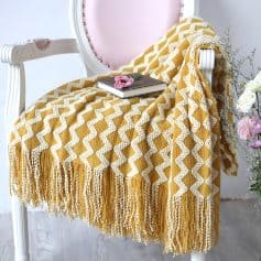 Mustard yellow and white knitted throw blanket with fringe on a vintage pink and white chair, with a book and pink flower on top, creating a cozy and stylish setting for home or bedroom decor.