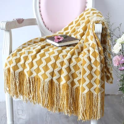 Mustard yellow and white knitted throw blanket with fringe on a vintage pink and white chair, with a book and pink flower on top, creating a cozy and stylish setting for home or bedroom decor.