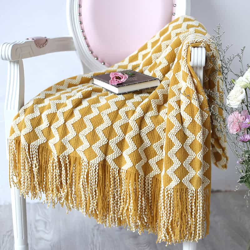 Mustard yellow and white knitted throw blanket with fringe on a vintage pink and white chair, with a book and pink flower on top, creating a cozy and stylish setting for home or bedroom decor.