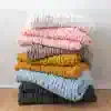 Luxurious chunky knit blankets stacked in pastel pink, cream, grey, mustard, blue, and charcoal, perfect for cozy home decor and elegant bedroom aesthetics.