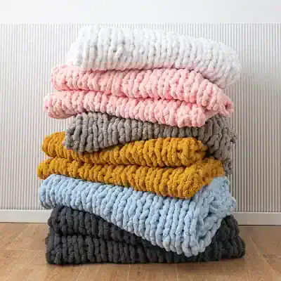 Luxurious chunky knit blankets stacked in pastel pink, cream, grey, mustard, blue, and charcoal, perfect for cozy home decor and elegant bedroom aesthetics.