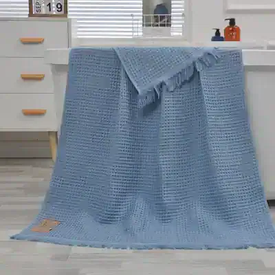 Blue waffle weave bath towel on white countertop with soap dispensers and window blinds.