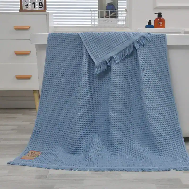 Blue waffle weave bath towel on white countertop with soap dispensers and window blinds.