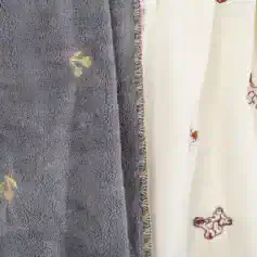 Soft plush gray towel with embroidered gold floral accents, next to cream fabric with teddy bear appliqué, ideal for cozy home or spa use.