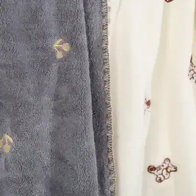 Soft plush gray towel with embroidered gold floral accents, next to cream fabric with teddy bear appliqué, ideal for cozy home or spa use.