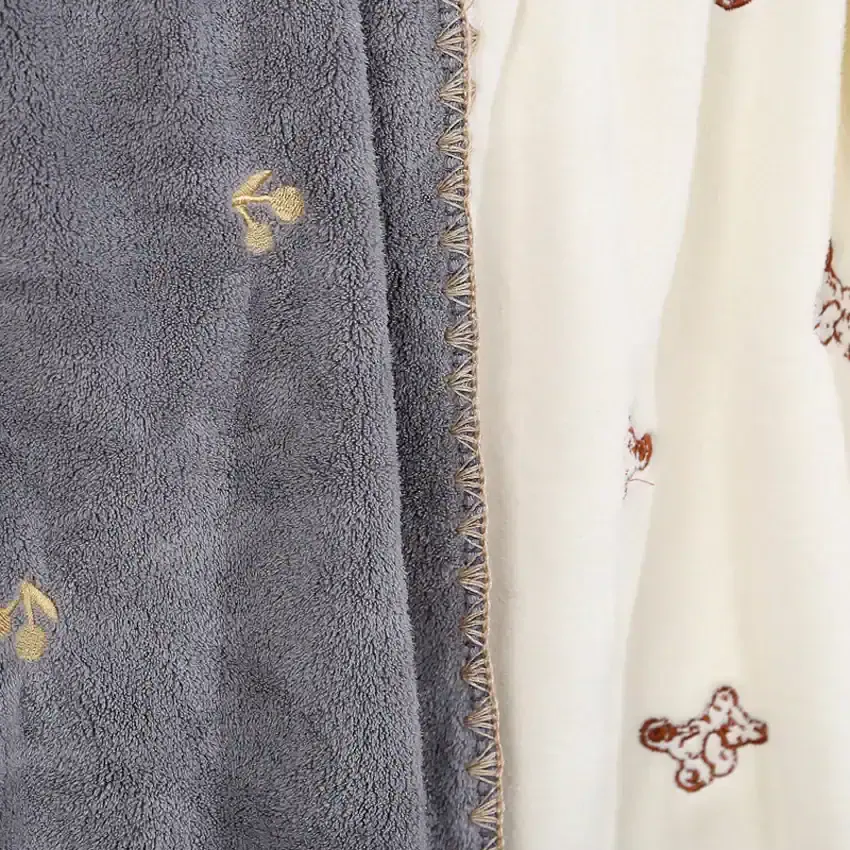 Soft plush gray towel with embroidered gold floral accents, next to cream fabric with teddy bear appliqué, ideal for cozy home or spa use.